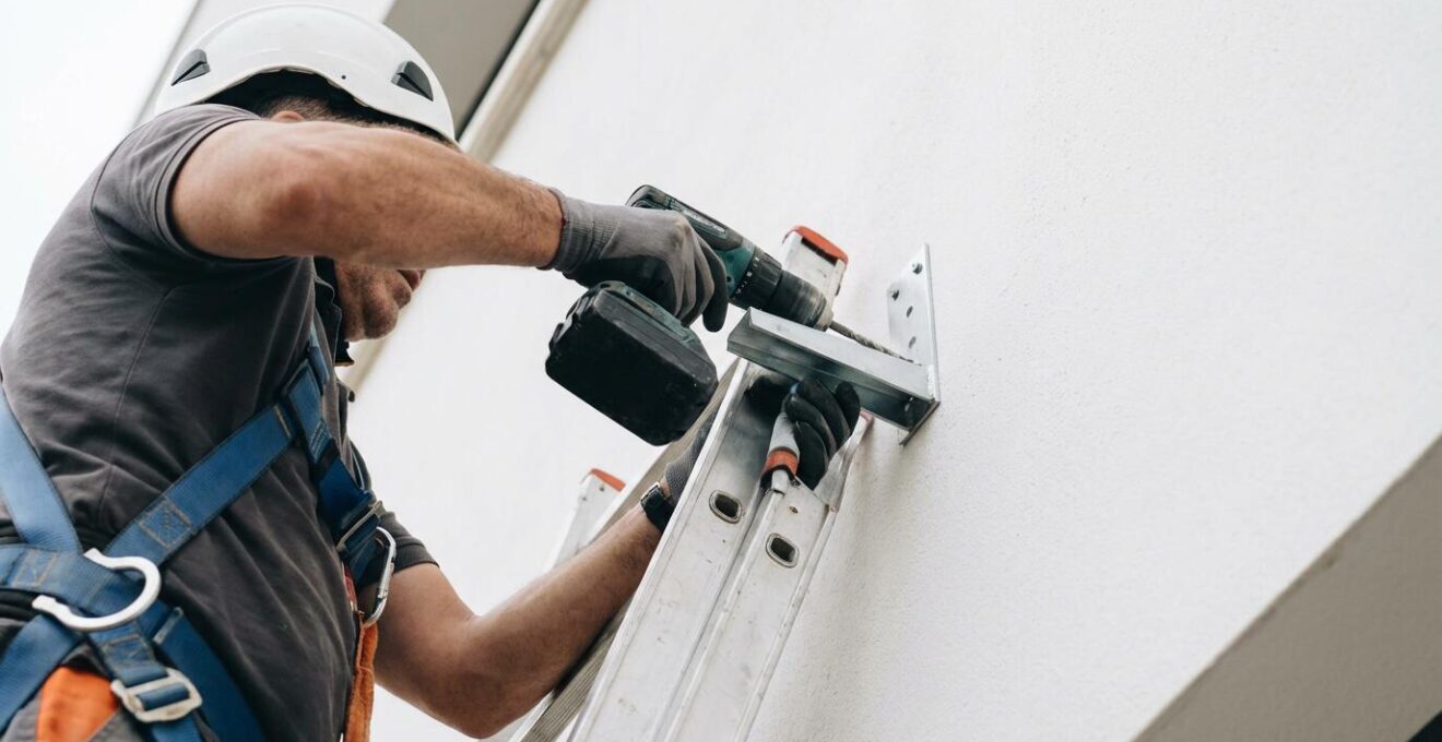 Instalador profesional sobre escalera fijando soporte de toldo cofre en fachada blanca
